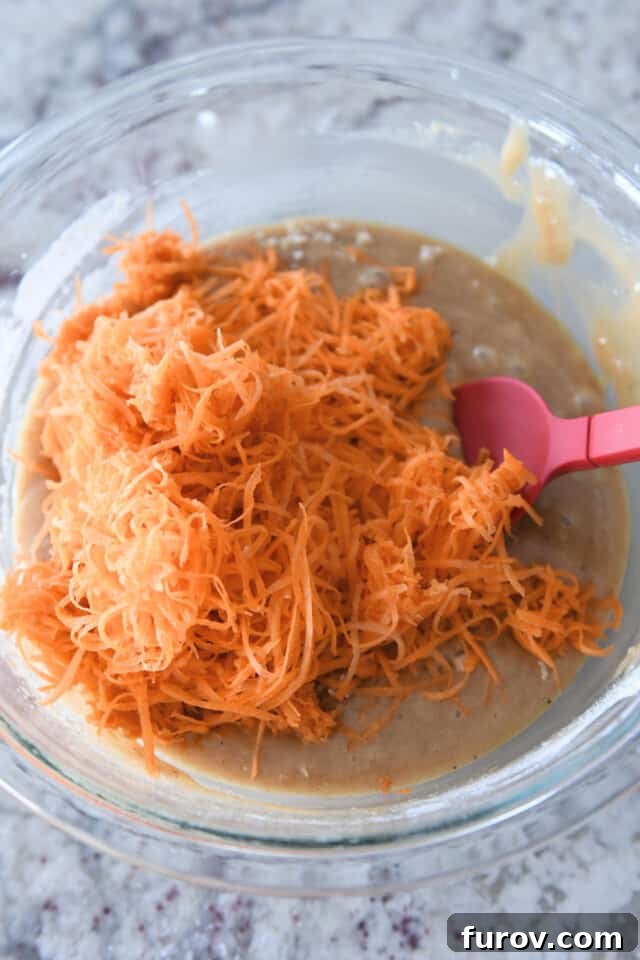 Freshly shredded carrots, vibrant orange in color, mixed into a golden cake batter in a clear glass bowl, illustrating the final step before baking.