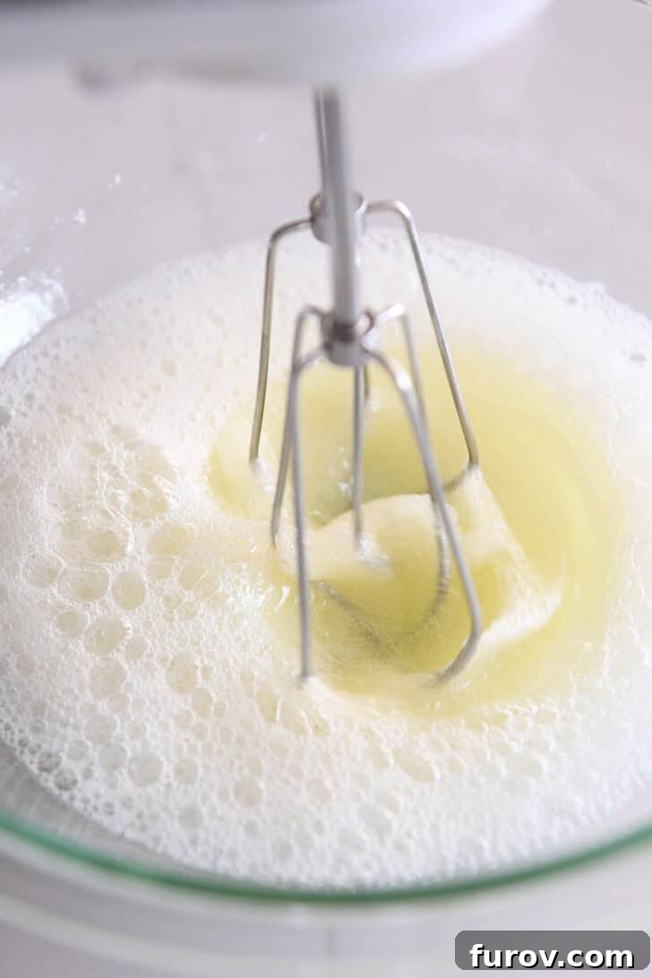 A frothy egg white mixture being whipped with an electric mixer in a clear glass bowl, illustrating the initial stages of meringue preparation.