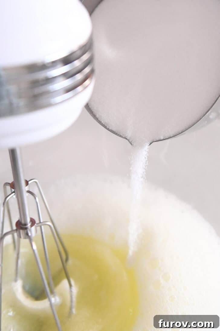 A metal measuring cup pouring granulated sugar into a glass bowl where egg whites are being whipped, demonstrating the gradual addition of sugar for stability.