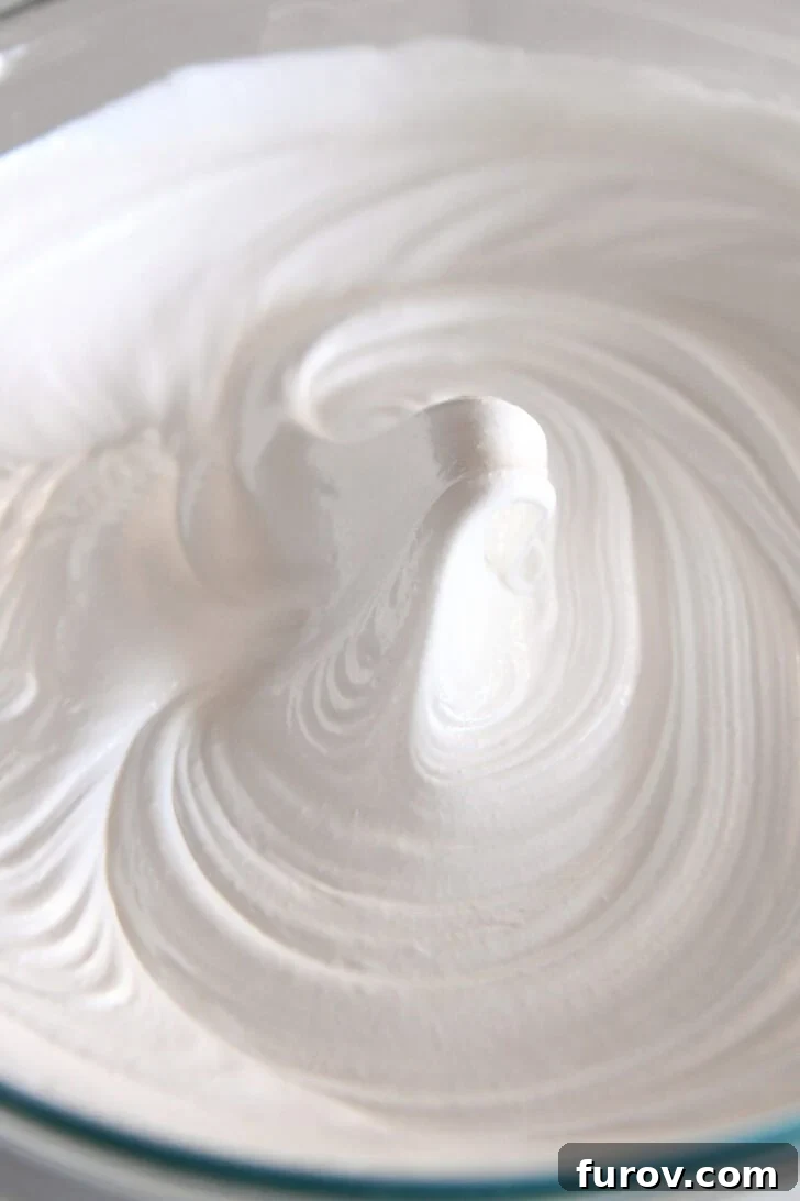 A glass bowl of perfectly whipped egg whites forming a stiff peak in the center, indicating the meringue is ready for baking and has achieved proper texture.