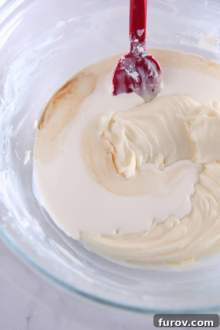 A glass bowl with a red spatula stirring a rich, creamy cream cheese mixture, showcasing the smooth, luscious texture of the cheesecake layer before it's whipped with cream.