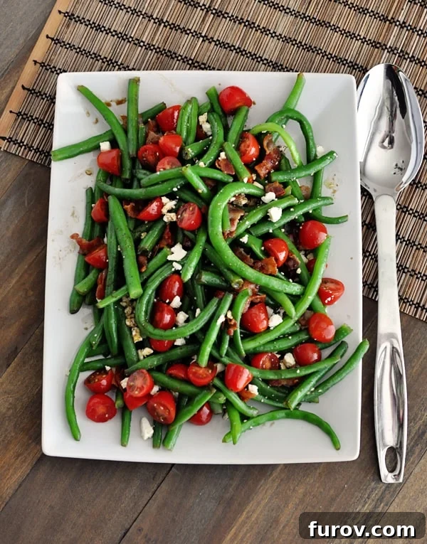 A top view of a white platter full of green beans, sliced cherry tomatoes, crumbled feta, and crispy bacon sprinkled throughout.