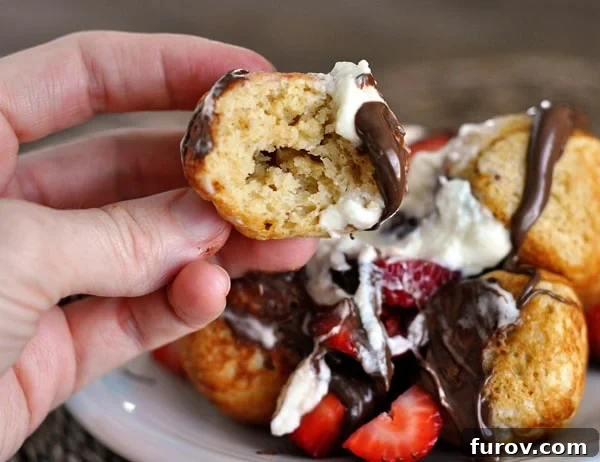 A half eaten ebelskiver in front of a plate of ebelskivers loaded with chocolate, strawberries, and whipped cream. A close-up of the delicious fillings and toppings.