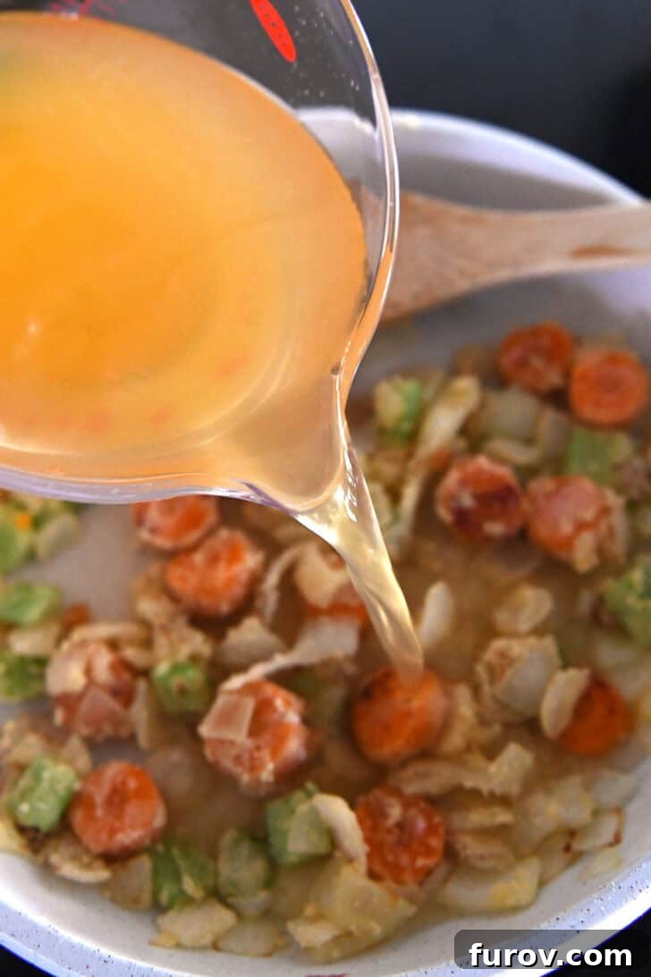 Pouring broth into white skillet with carrots, celery and onions.