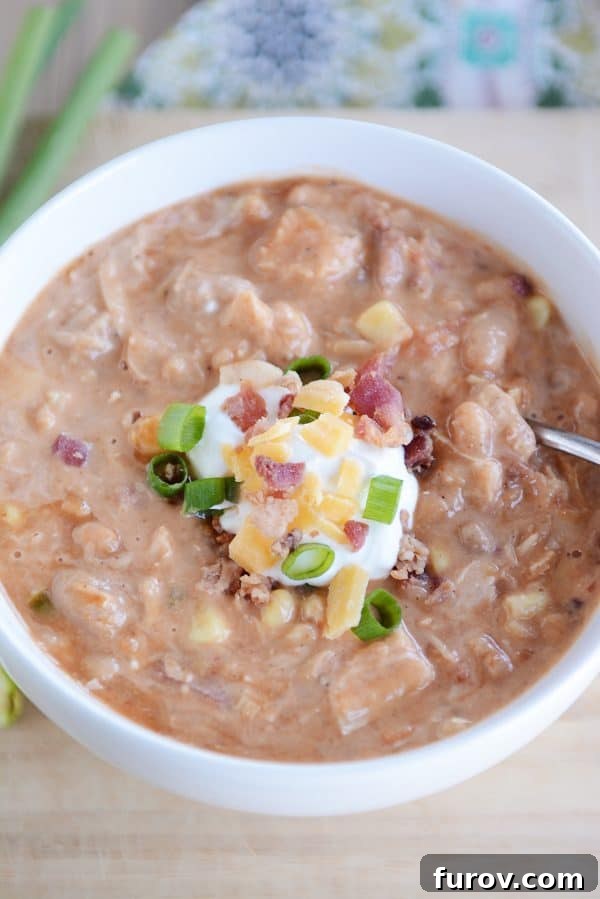 An overhead shot of a cozy bowl of jalapeño popper white bean chicken chili, perfectly garnished with a dollop of sour cream, a sprinkle of shredded cheese, and fresh green chives.