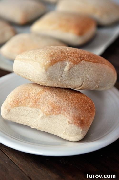 Mastering Chewy Italian Rolls 2 Two golden brown rectangular shaped italian rolls stacked on top of each other on a white plate, with the rest of the rolls on a cookie sheet in the background.