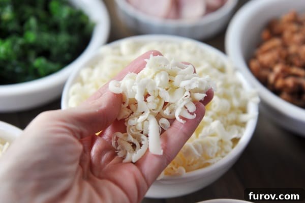 A person's hand sprinkling freshly shredded cheese over a bowl of cheese, with other pizza toppings visible.
