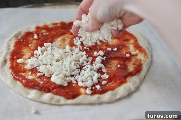 An uncooked pizza crust with red sauce, being sprinkled with freshly shredded cheese.