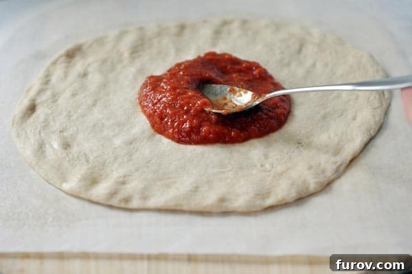An uncooked pizza crust with a vibrant red sauce being spread evenly over it.