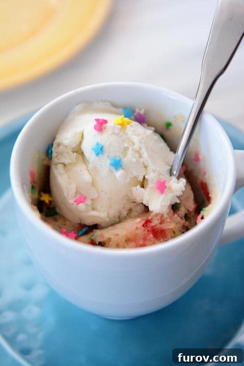 A vibrant funfetti mug cake, freshly cooked in a white mug, topped with a generous scoop of vanilla ice cream and colorful sprinkles, ready to be enjoyed with a spoon.
