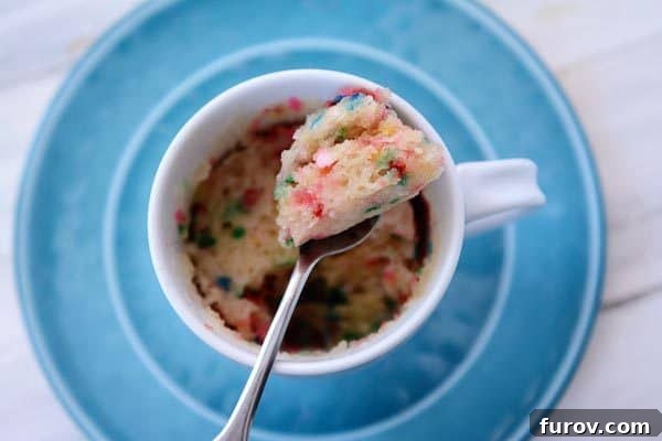 A close-up, top-down view of a spoon digging into the warm, fluffy interior of a vanilla funfetti mug cake, highlighting its delightful texture.