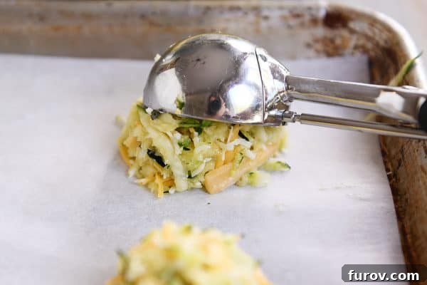 A cookie scoop depositing a perfectly sized mound of uncooked cheesy zucchini batter onto a parchment-lined baking sheet, demonstrating the initial shaping step.