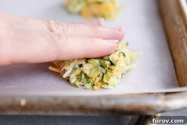 A hand gently patting down an uncooked cheesy zucchini bite on a cookie sheet, flattening it into a disc shape for even baking and crispiness.