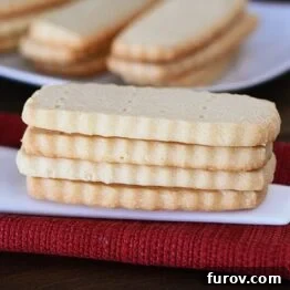 A stack of golden-brown shortbread cookies with more cookies arranged on a white plate in the background.