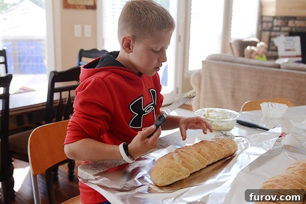 A young boy meticulously spreading a creamy cheese mixture onto a golden-brown loaf of French bread.