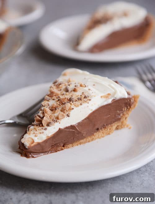 Delicious slice of homemade chocolate cream pudding pie with a golden graham cracker crust and fluffy whipped cream topping on a white plate.