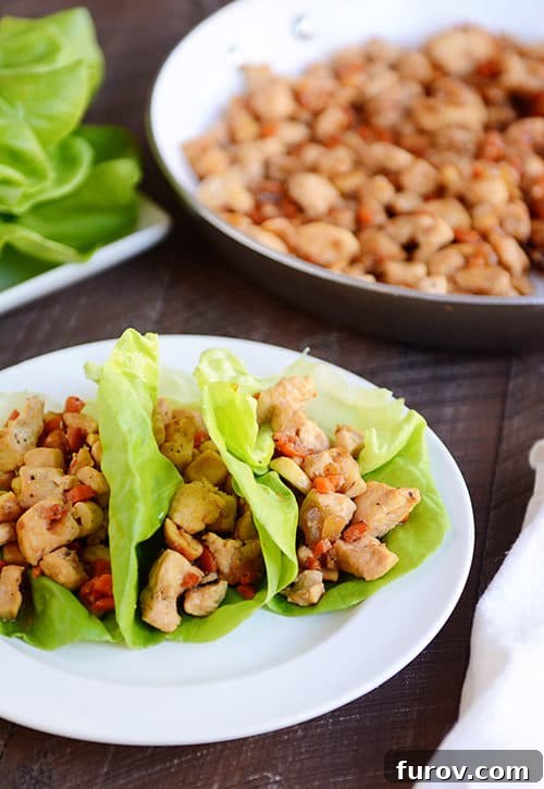 Three cashew chicken lettuce wraps on a white plate with a skillet of chicken mixture behind it.