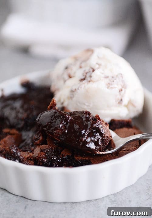 A white ramekin with a generous serving of brownie pudding, accompanied by a scoop of ice cream, with a spoon ready for a bite.