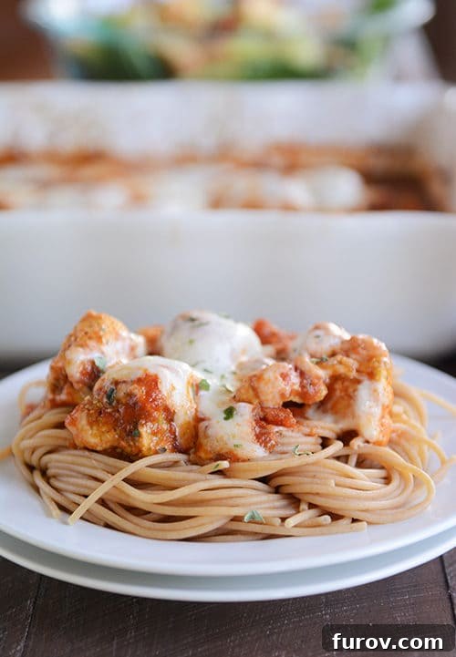 Cheesy Pesto Marinara Chicken Bake 2 A white plate of cooked spaghetti topped with cheesy marinara chicken, garnished with fresh basil.