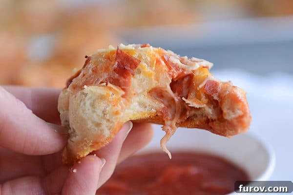 A close-up of a pepperoni pizza roll with a bite taken out, revealing the layers of dough, melted cheese, and pepperoni, held over a bowl of marinara sauce.