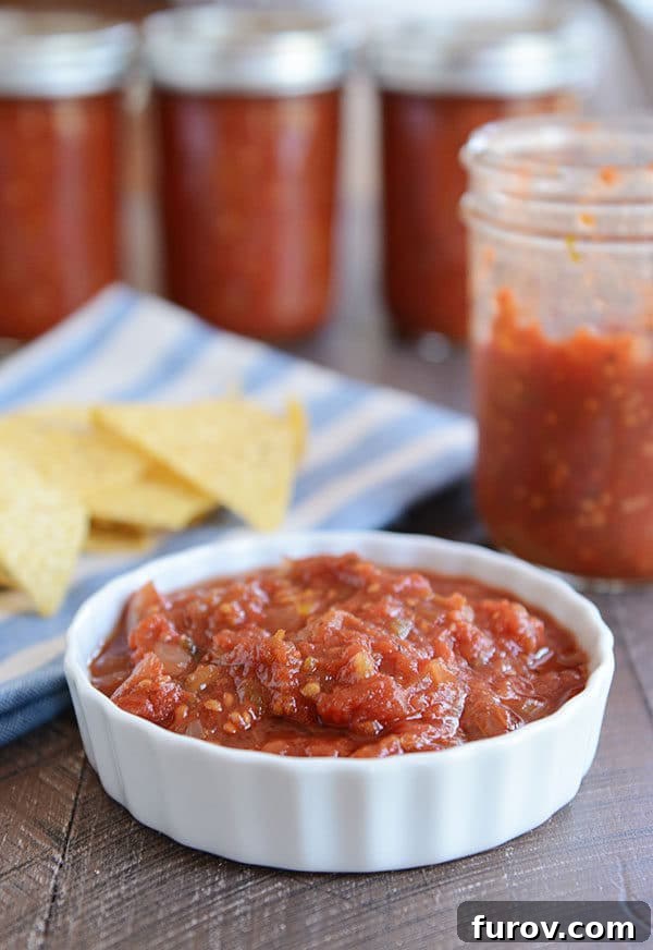 Perfectly Crafted Homemade Salsa 2 A small ramekin filled with freshly made homemade salsa, with a half-empty jar of the same salsa and a pile of crispy tortilla chips in the background.