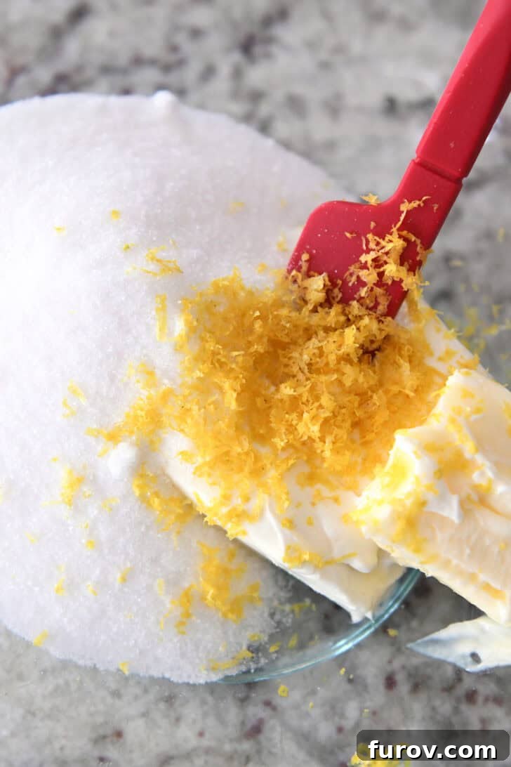 Top down view of a glass bowl with sugar, lemon zest, cream cheese, and a red spatula.