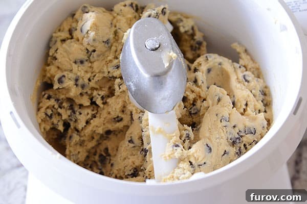 A perfectly mixed, large batch of chocolate chip cookie dough rotating within a white Bosch stand mixer bowl, ready for baking.