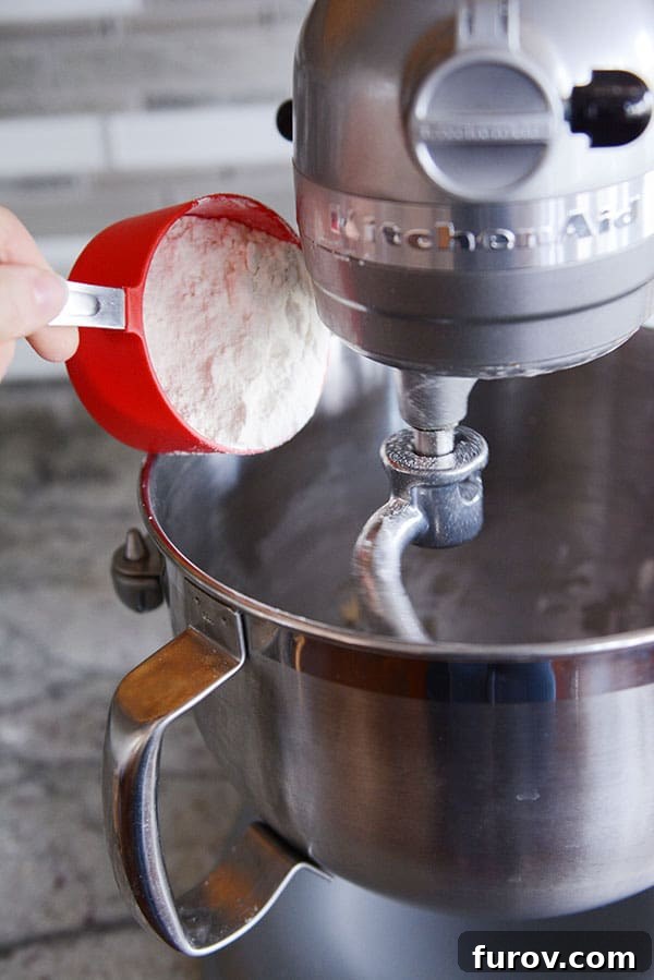White flour being carefully poured from a measuring cup into the bowl of a KitchenAid stand mixer, illustrating the process of adding dry ingredients.