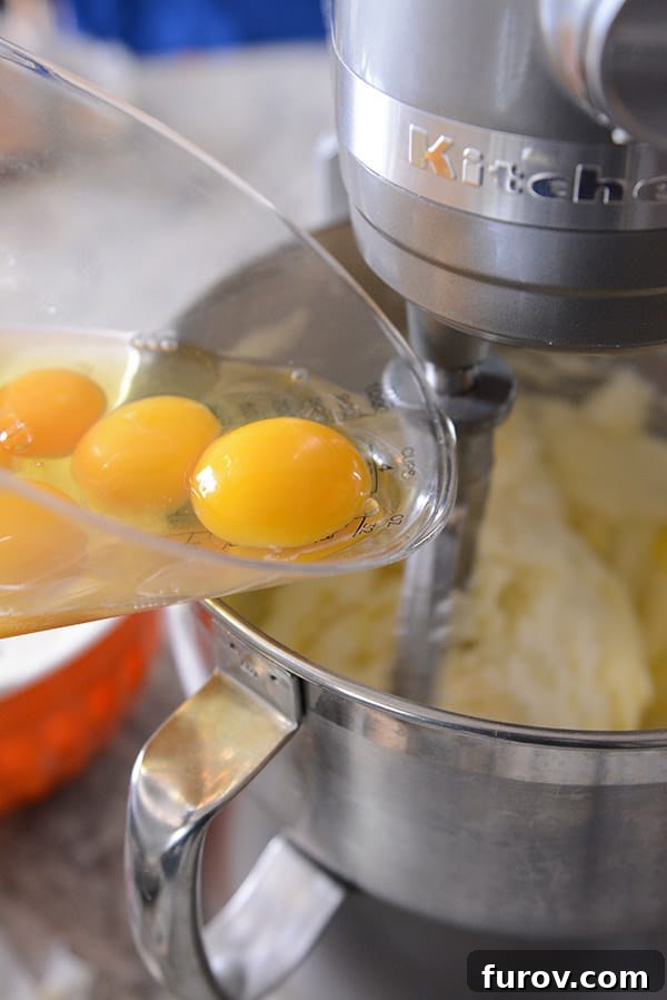Raw eggs being gently poured from a measuring cup into the mixing bowl of a KitchenAid stand mixer, demonstrating liquid ingredient addition.