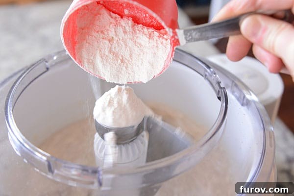 White flour being swiftly poured into the large, open bowl of a Bosch stand mixer, showcasing its ease of adding ingredients.
