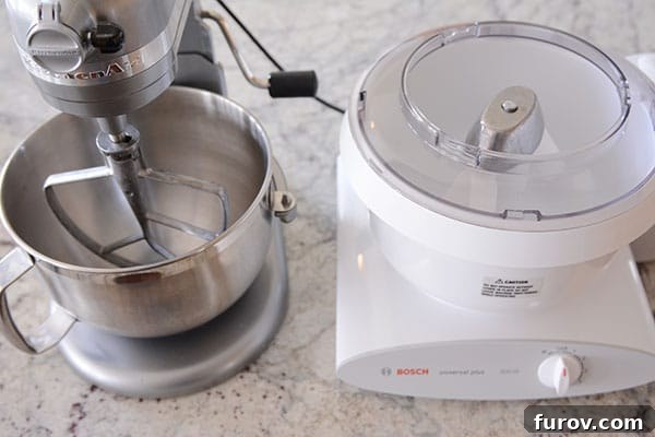 A KitchenAid stand mixer and a Bosch Universal stand mixer displayed side-by-side on a clean kitchen countertop, highlighting their distinct designs.