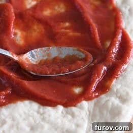 A metal spoon spreading pizza sauce on flattened, uncooked pizza dough.