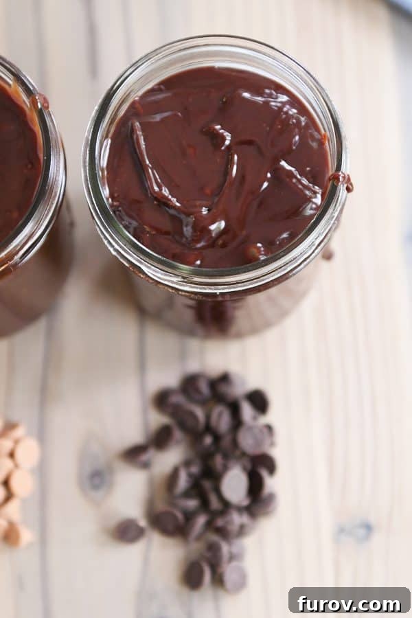 Rich chocolate 5-minute hot fudge sauce stored in a glass jar, with scattered chocolate chips on a kitchen counter, showcasing its decadent appearance.