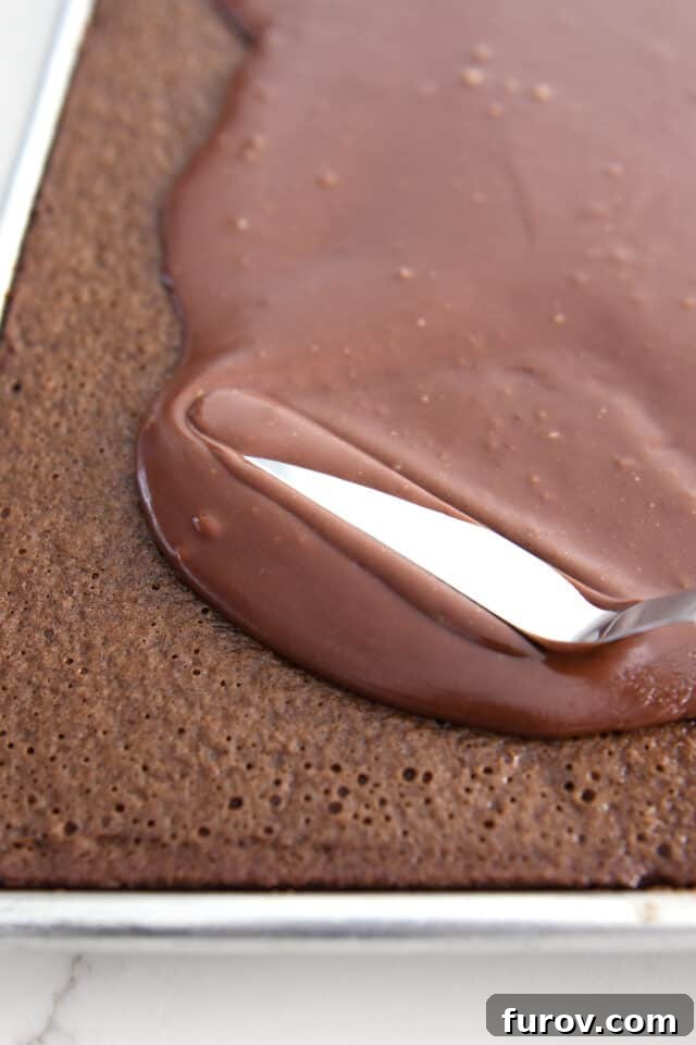 Using offset spatula to spread thin frosting over chocolate cake in sheet pan. Evenly distributing the warm chocolate glaze.