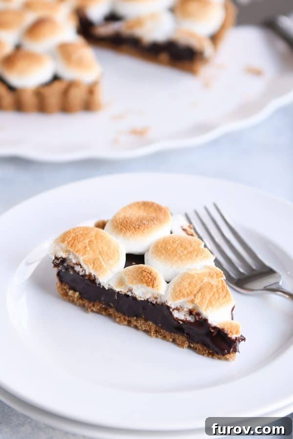 A piece of s'mores pie on a white plate with a fork, showing the perfect layers and toasted marshmallow.