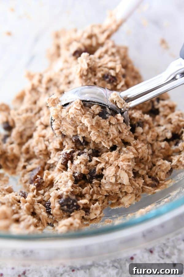 An ice cream scoop delicately scooping out a portion of rich oatmeal cookie dough from a large glass bowl.