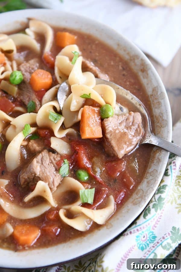 Top down view of slow cooker beef vegetable noodle soup with spoon in gray ceramic bowl.