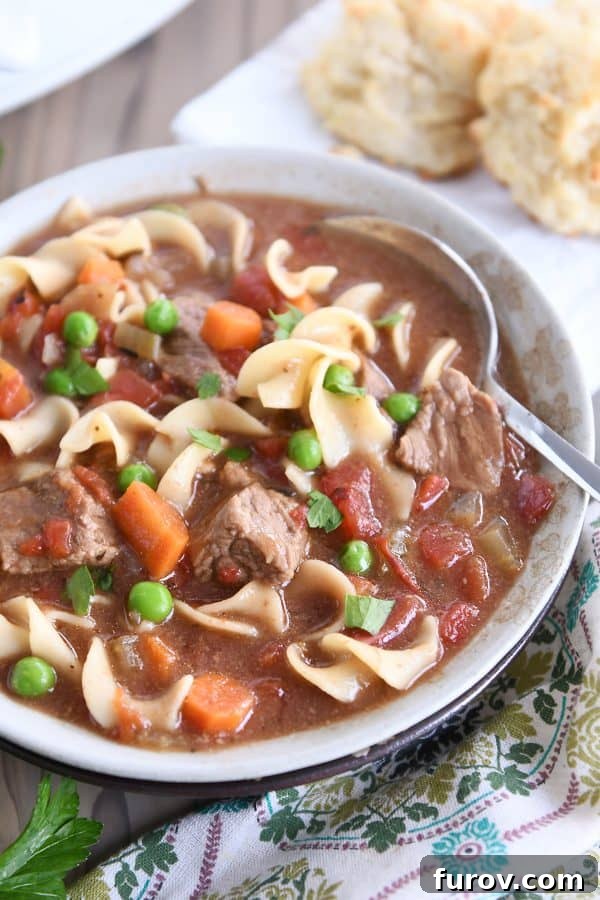 Slow cooker beef vegetable noodle soup with spoon in gray ceramic bowl.