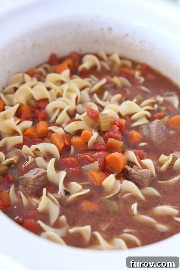 White slow cooker filled with beef vegetable noodle soup.