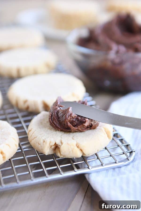 A hand gently spreading rich, fluffy chocolate frosting onto a soft peanut butter sugar cookie, creating a perfect dessert.