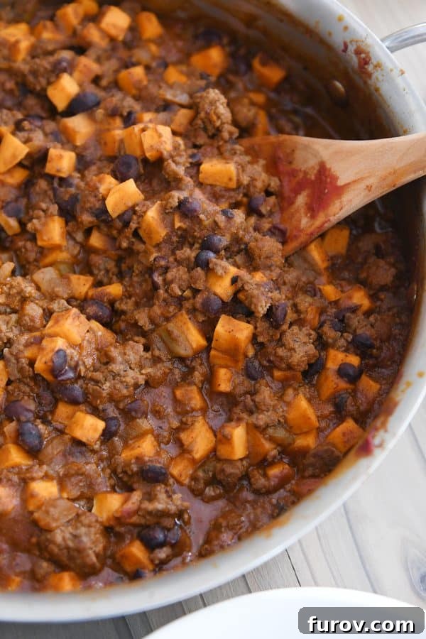 A wooden spoon rests in a cast-iron skillet filled with the rich, simmering sweet potato and black bean burrito mixture, highlighting its hearty texture and vibrant colors.