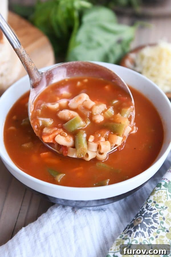 A ladle pouring rich, classic minestrone soup into a white ceramic bowl.