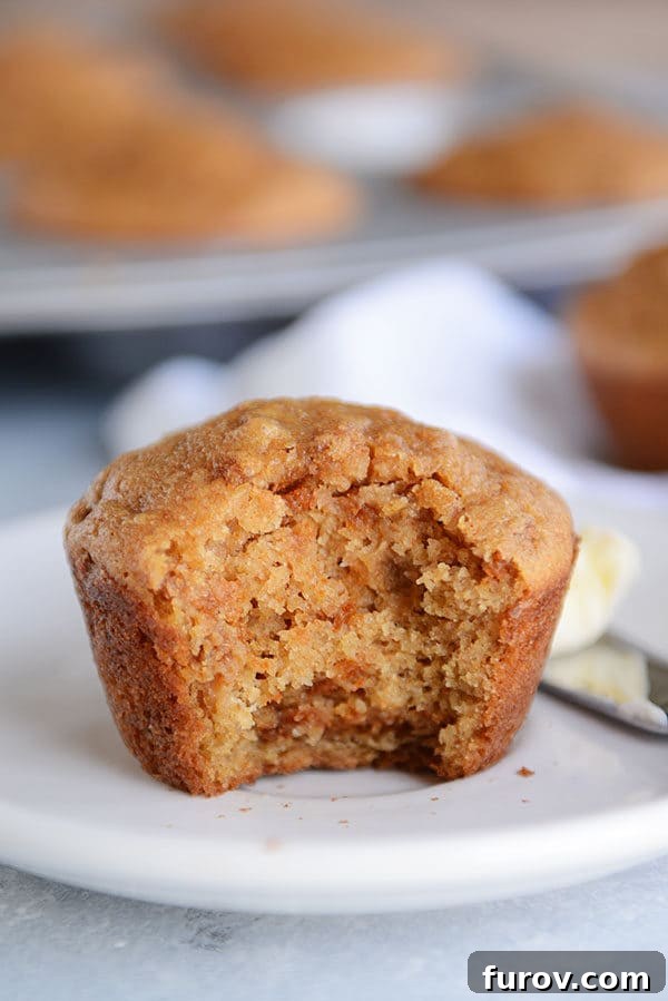 A cooked bran muffin with a bite taken out on a white plate and a pat of butter on a knife next to the muffin.