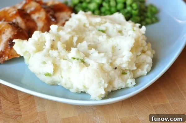 A generous scoop of creamy baked garlic mashed potatoes on a plate, with vibrant green peas and a slice of perfectly cooked pork tenderloin in the background, showcasing its golden-brown topping.
