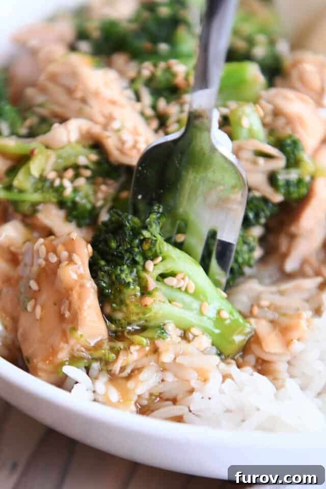 White bowl with rice, broccoli, chicken, sesame seeds and fork. A generously saucy and satisfying Chinese chicken and broccoli meal.