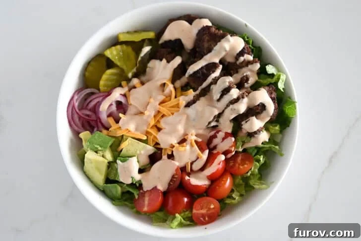 Top down view of lettuce, ground beef, tomatoes, avocados, pickled onions, pickles, shredded cheese and creamy sauce in white bowl.