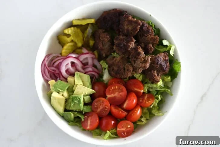 Top down view of lettuce, ground beef, tomatoes, avocados, pickled onions, pickles in white bowl.