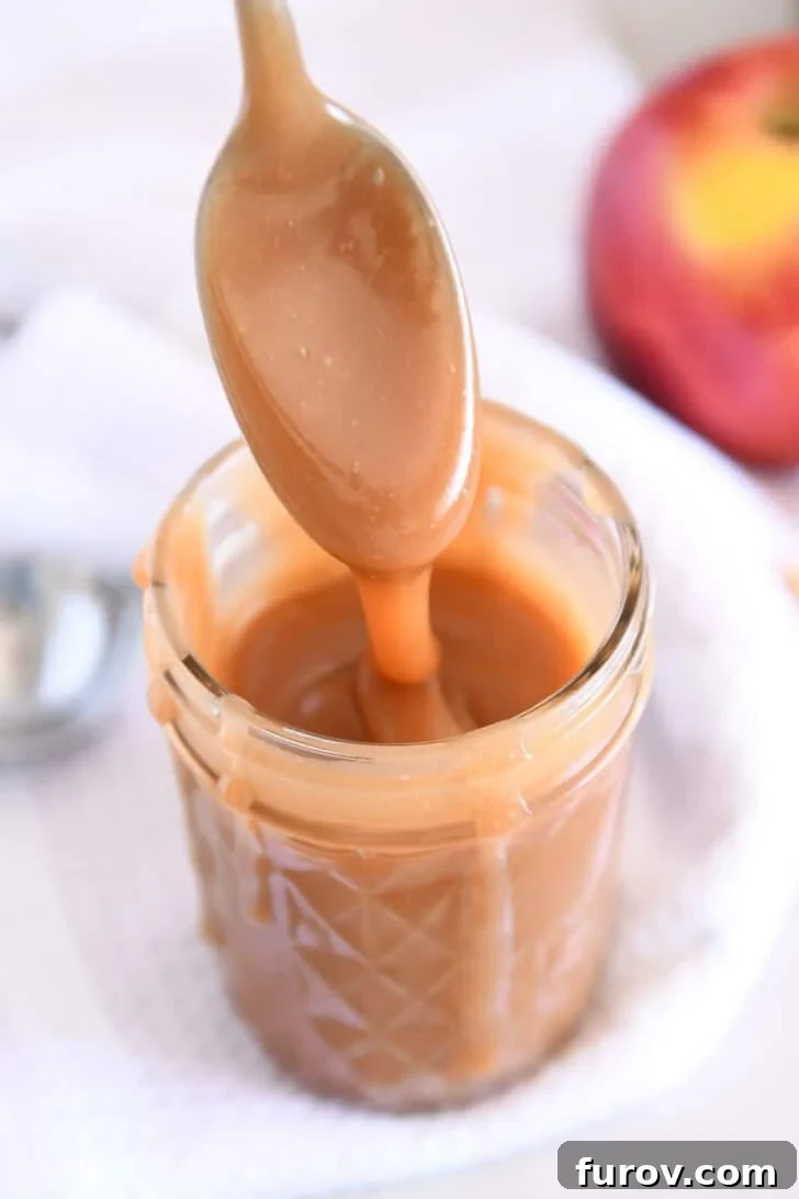 Spoon drizzling caramel sauce into glass jar with apple in the background.