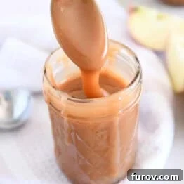 Spoon drizzling caramel sauce into glass jar with apple in the background.
