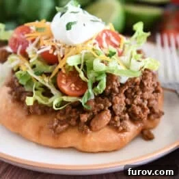 Piece of fry bread on cream plate topped with taco meat, lettuce, tomatoes, cheese, and sour cream.